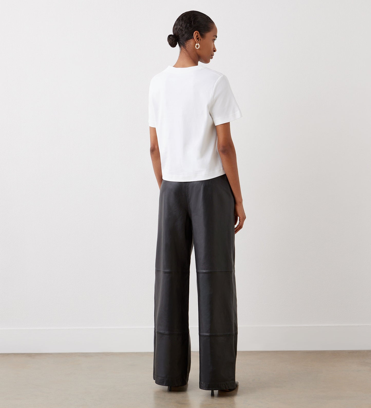 A woman with her hair pulled back and hoop earrings stands indoors facing away, wearing Theresa Black High Waisted Wide Leg Leather Trousers, a white short-sleeve top, and black shoes against a plain white wall and light-colored floor.