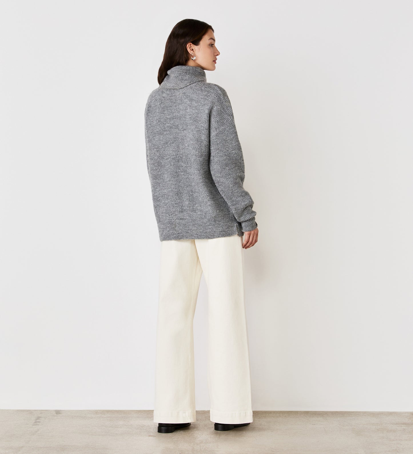 A woman faces away from the camera wearing the Tillie Grey Marle Roll Neck Jumper, paired with white wide-leg pants with side splits, black shoes, and hoop earrings, set against a plain white wall and concrete floor.