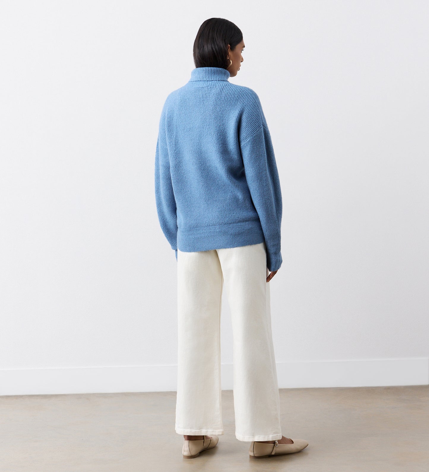 A person with short dark hair faces away, wearing a Tillie Blue Roll Neck Jumper, wide-leg white pants, and beige shoes, standing against a plain white wall and light-colored floor.