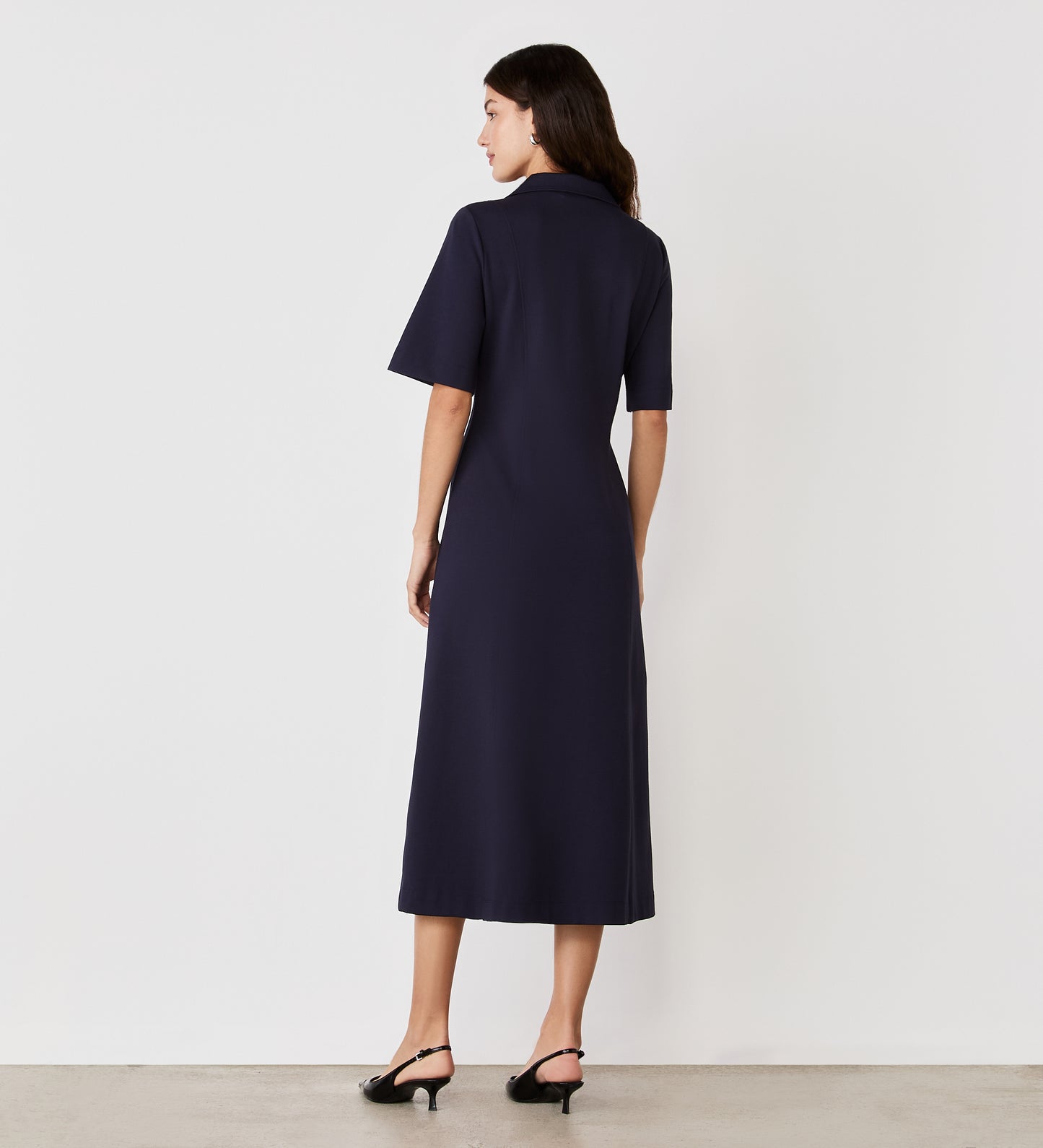 A woman stands facing away, wearing the Trinity Navy Ponte Jersey Midi Dress with short sleeves and black heeled sandals, against a plain white background.