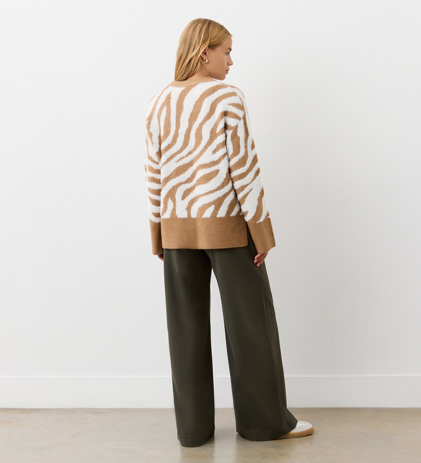 A person stands facing away, wearing the Tully Tan Zebra Knitted Jumper with brown cuffs and hem, paired with wide-leg dark olive pants and white sneakers, against a plain white wall.