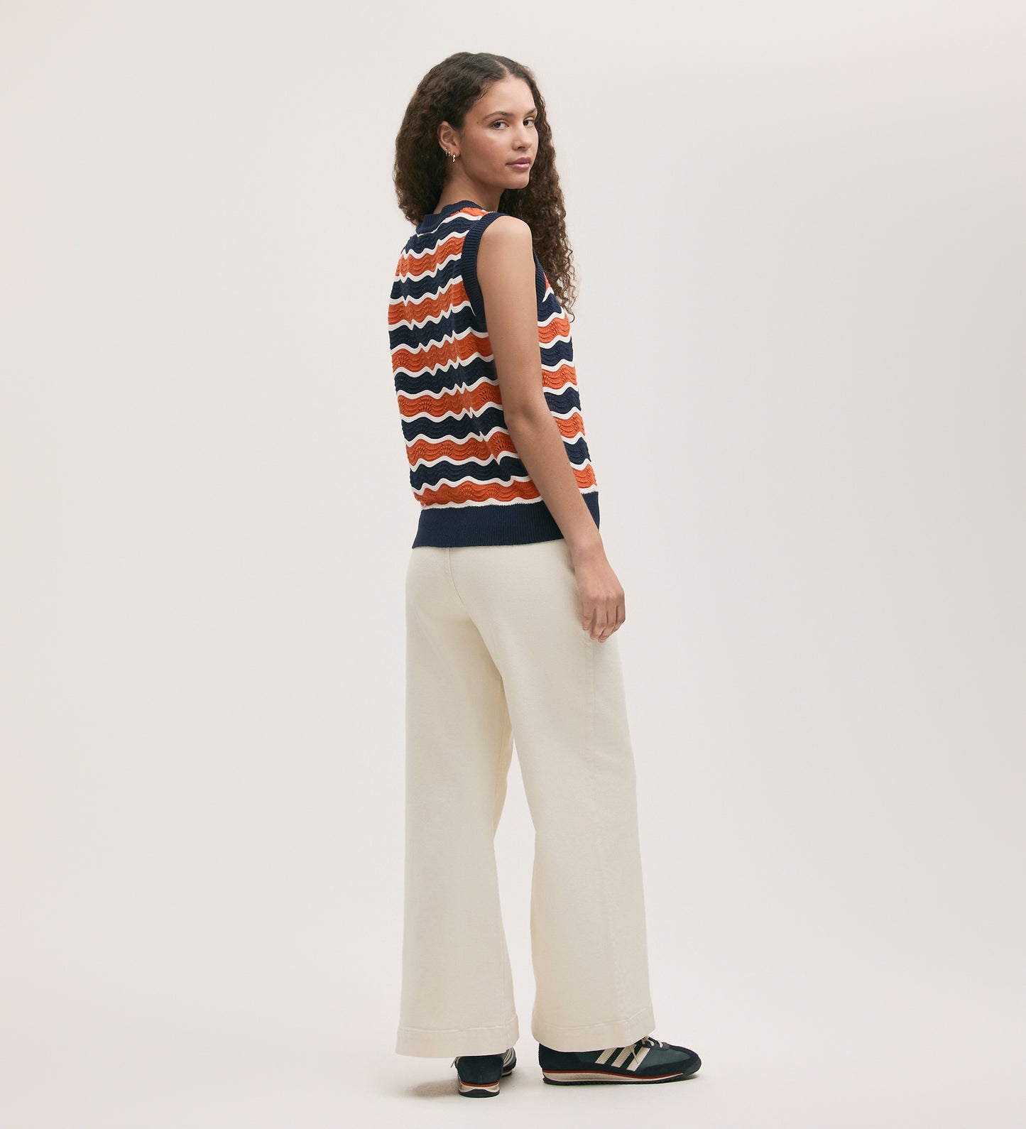 A woman with long curly hair wears the Taylor Orange Stripe Crochet Vest, wide-leg white pants, and black sneakers, standing and looking over her shoulder against a plain light background.