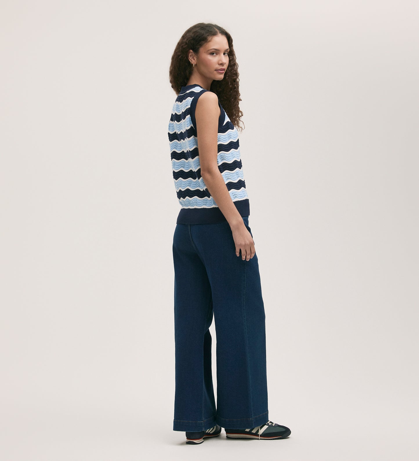 A woman with curly hair stands sideways against a light background, wearing the Taylor Blue Stripe Crochet Vest in navy and white, paired with wide-leg dark blue jeans and black shoes.