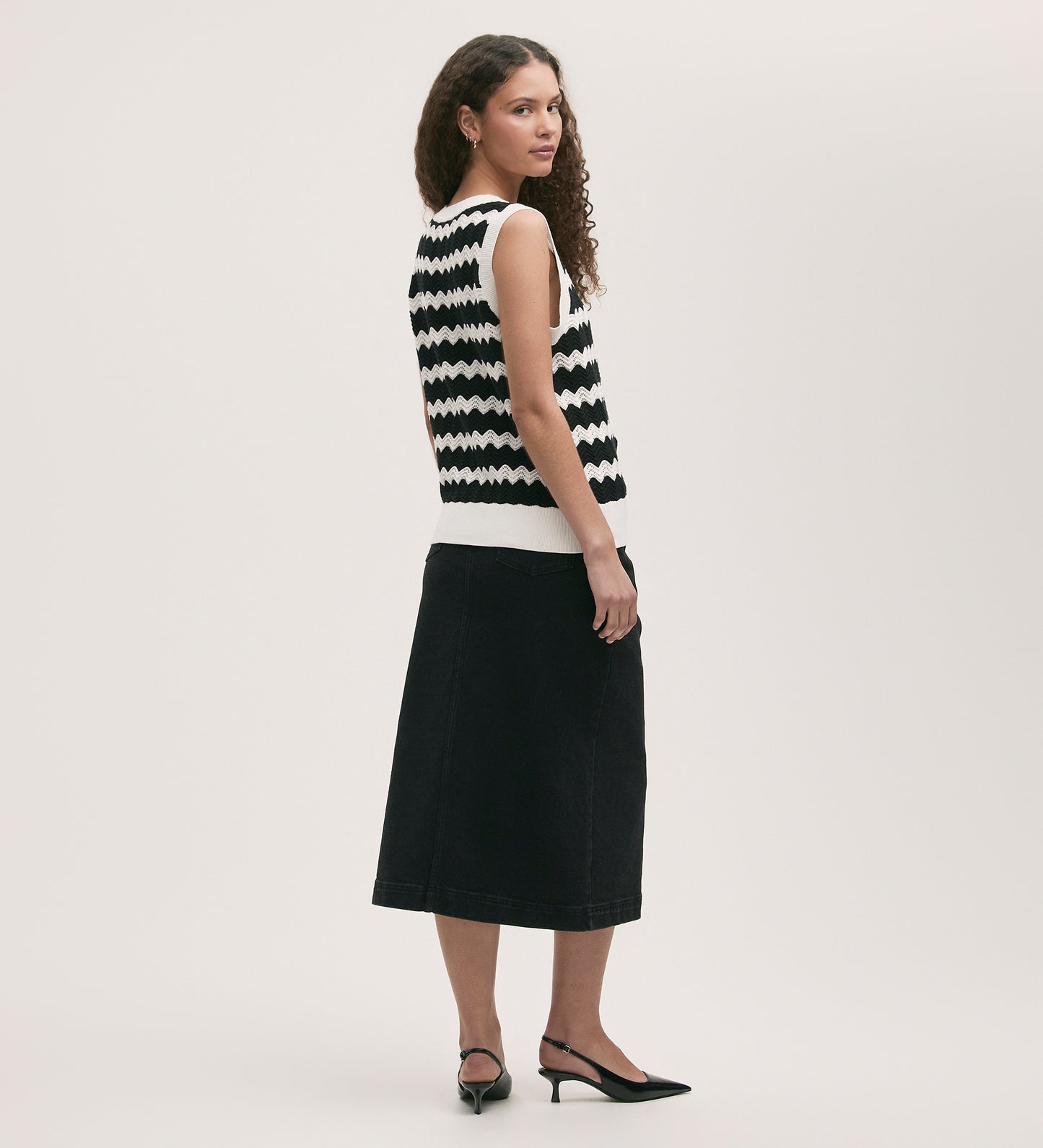 A woman stands sideways, looking over her shoulder, wearing the Tessa Black Stripe Crochet Vest with a black midi skirt and black slingback heels against a plain light-colored background.