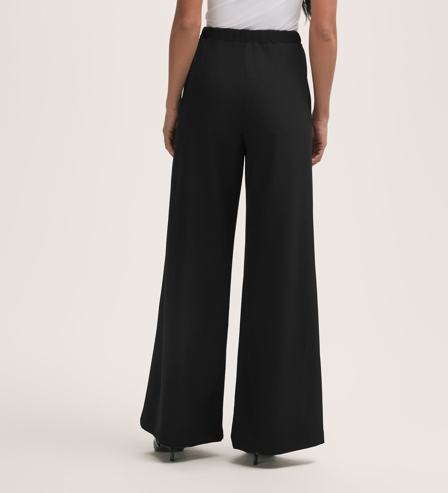 A woman stands with her back to the camera, wearing Nat Black Crepe Wide Leg Trousers with a relaxed, high-waisted fit and a white top. She pairs them with black heels against a plain light beige background.