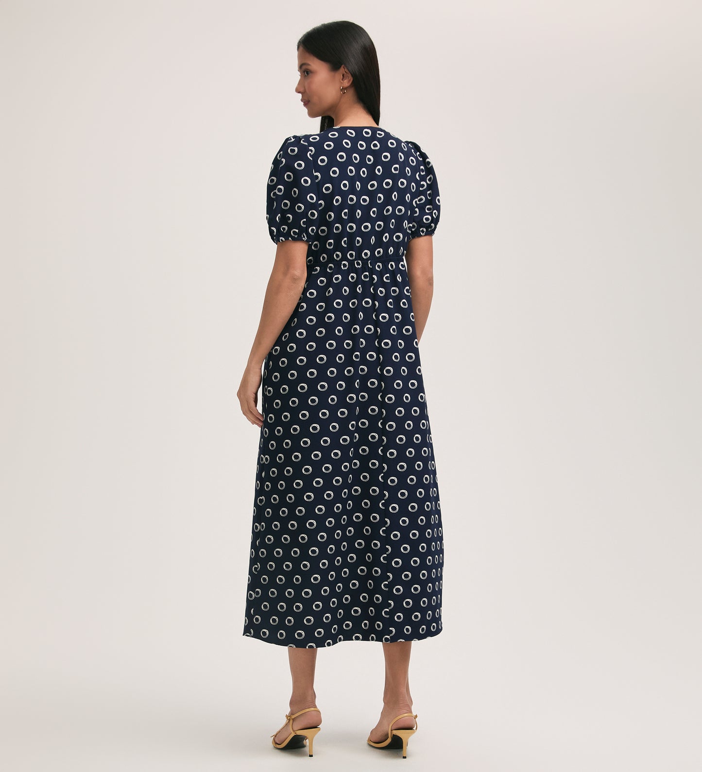 A woman faces away, wearing the Vittoria Navy Polka Dot Crepe Midi Dress with white spots and short puff sleeves, paired with tan heeled sandals. The crease-free fabric adds to her chic look against a light, plain background.