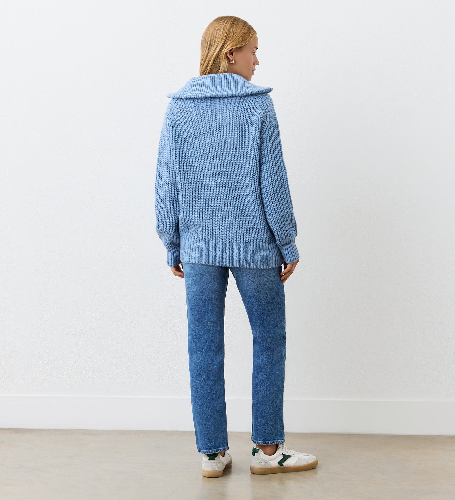 A woman with blonde hair stands by a white wall in a minimal setting, wearing the Wilma Denim Blue Half Zip Funnel Neck Jumper, blue jeans, and white sneakers with beige accents on a light wood floor.