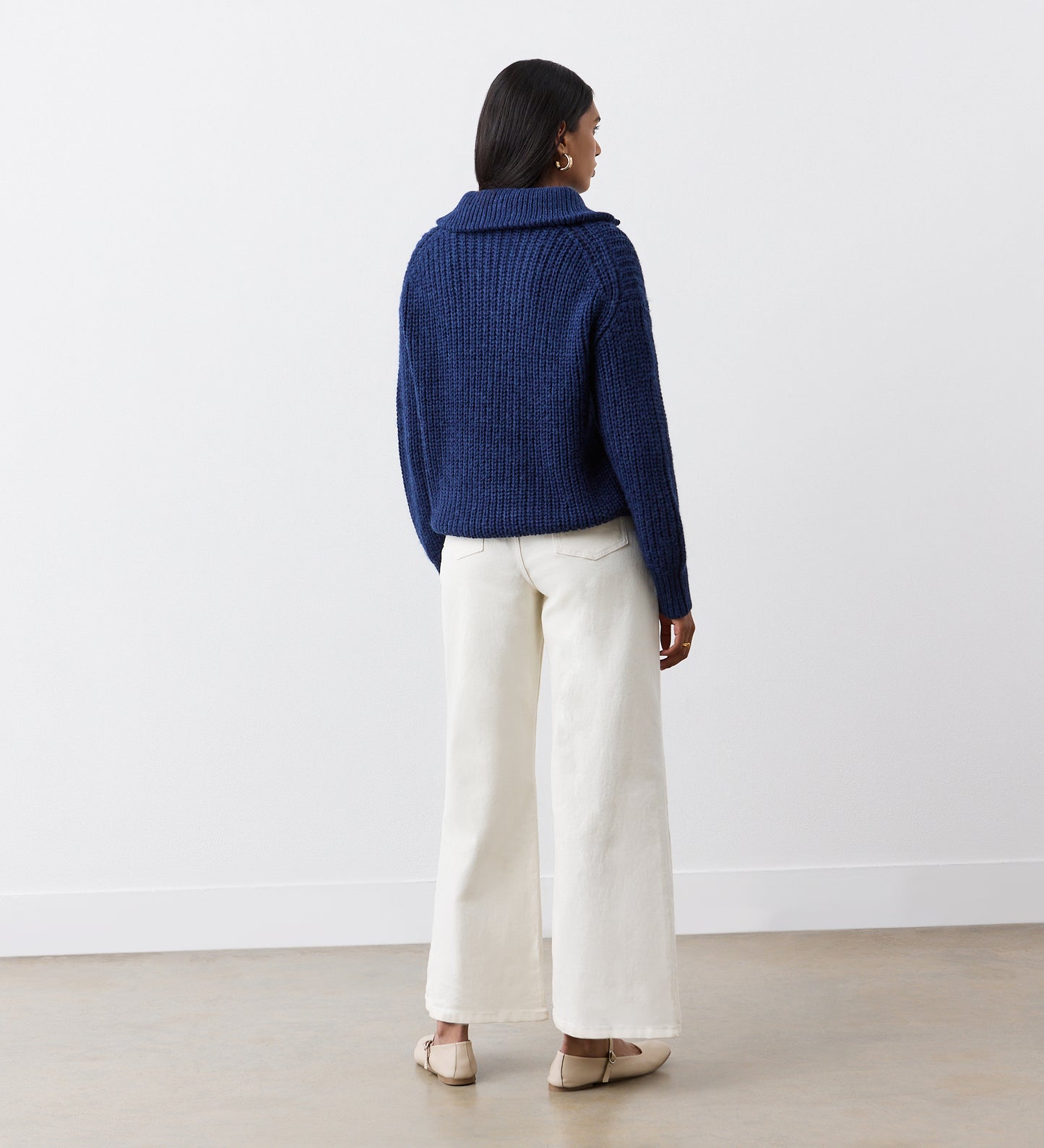 A woman faces away from the camera, wearing the Wilma Navy Half Zip Funnel Neck Jumper with long sleeves and wide-leg white pants, paired with beige flat shoes, against a plain white wall and light-colored floor.