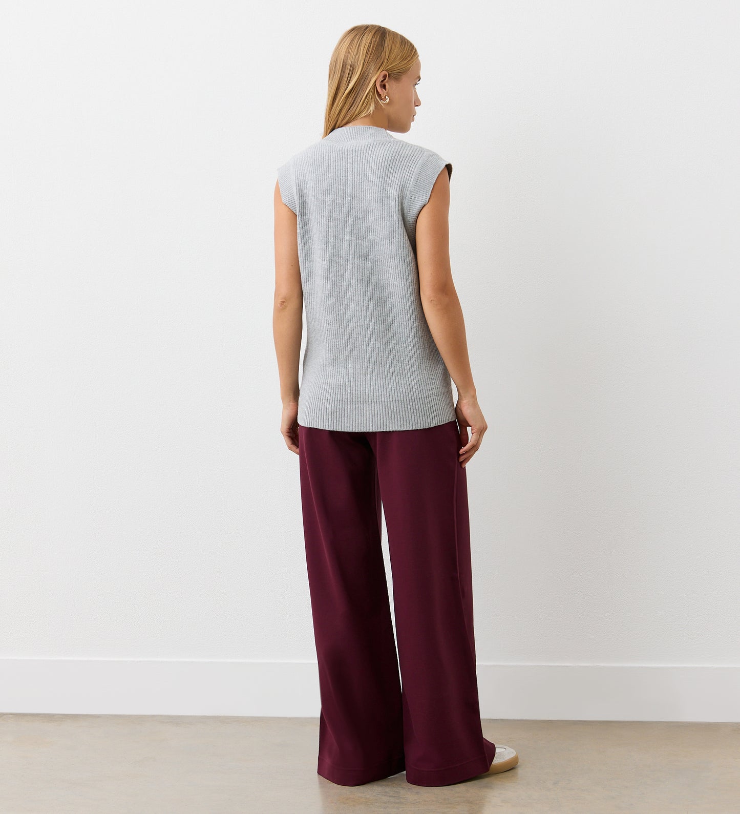 A woman with straight blonde hair, facing away, wears the Winnie Grey Marle Sleeveless Cardigan and loose burgundy pants with white sneakers, standing against a plain white wall and light-colored floor.