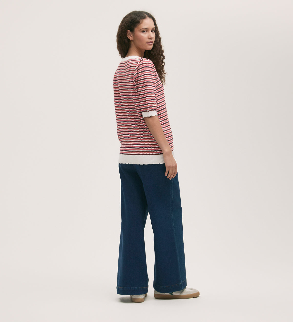 A woman with long curly hair wears the Willa Pink Stripe Cardigan with scallop ribbed details, paired with wide-leg blue jeans and beige shoes, standing slightly turned to the side against a plain light background.