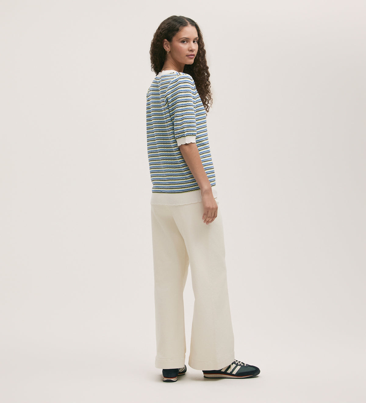 A woman with long curly hair stands sideways, looking back at the camera. She wears the Willa Blue Stripe Cardigan over wide-leg cream pants and navy sneakers with white stripes, set against a plain light background.