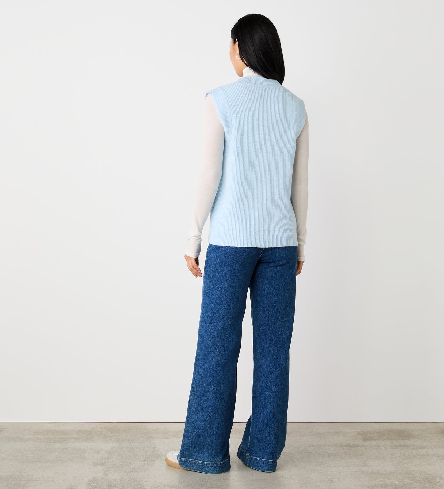 A woman with straight dark hair faces away, wearing the Winnie Pale Blue Sleeveless Cardigan over a long-sleeve white top and wide-leg blue jeans, standing against a plain white wall and light floor.