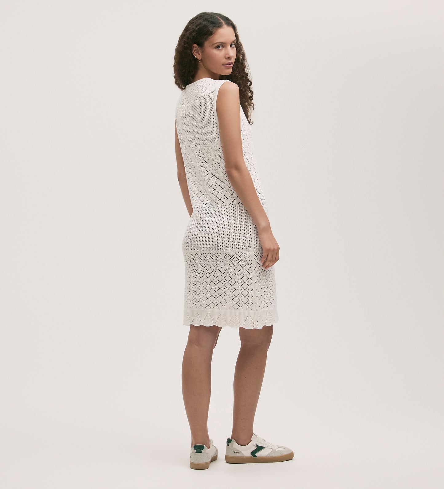 A woman with curly hair wears the Zendaya Ivory Crochet Knee Length Dress and white sneakers, standing at an angle and glancing over her shoulder against a plain light background.