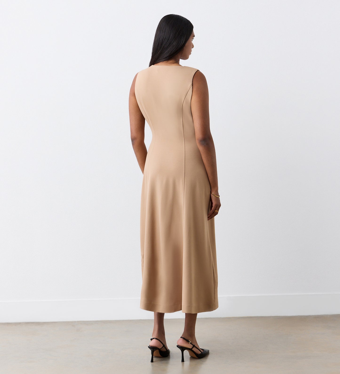 A woman with long dark hair, seen from behind, wears the Adele Camel Ponte Jersey Midi Dress with black heeled sandals, standing against a plain white wall on a concrete floor.