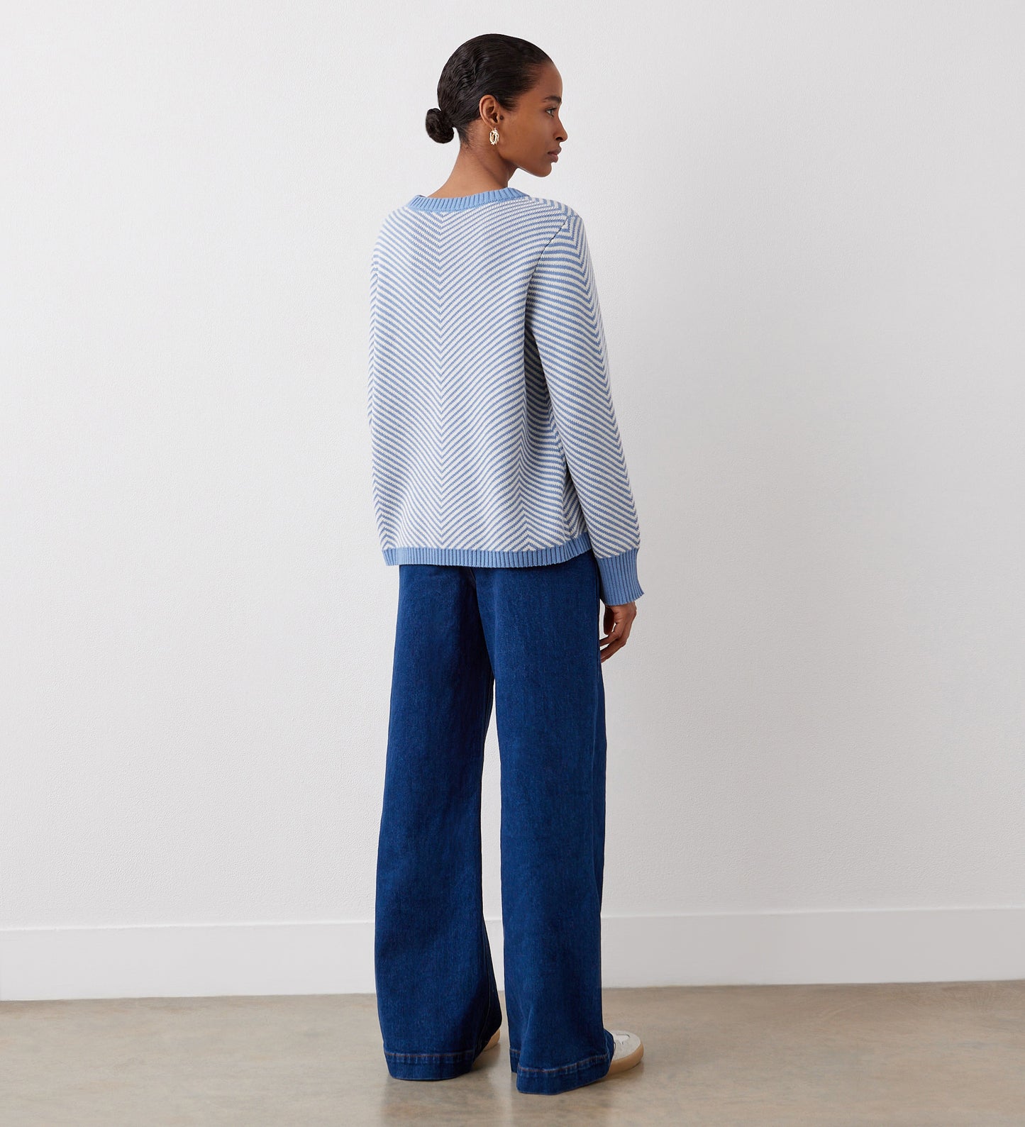 A woman with hair in a low bun faces away, wearing the Stella Blue Chevron Cotton Crew Neck Jumper and wide-leg blue jeans, in a minimal room with white walls and a brown floor.