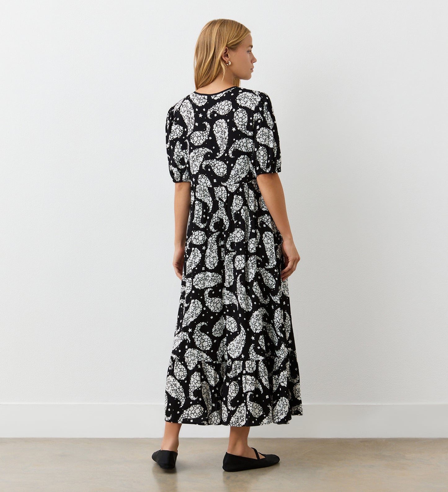 A woman with blonde hair stands sideways against a white wall, wearing the Cyra Black Paisley Crepe Tie Neck Dress, featuring a white floral and paisley pattern, tiered hem, and black flats on a light-colored floor.