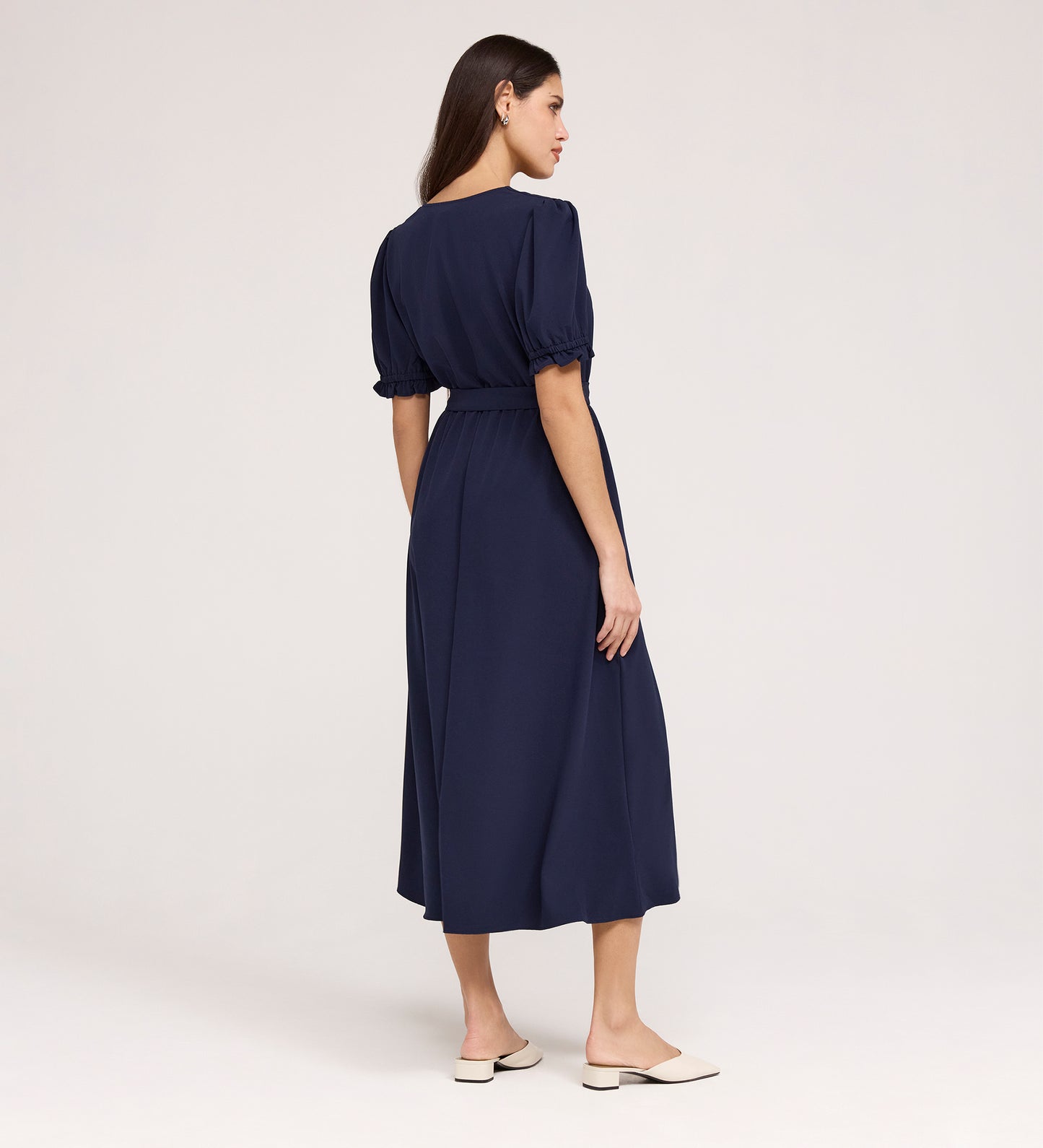 A woman with long brown hair, seen from behind, wears the Everleigh Navy Crepe Midi Dress featuring a faux wrap neckline and elastic waist. She pairs it with cream flats and stands against a plain light background.