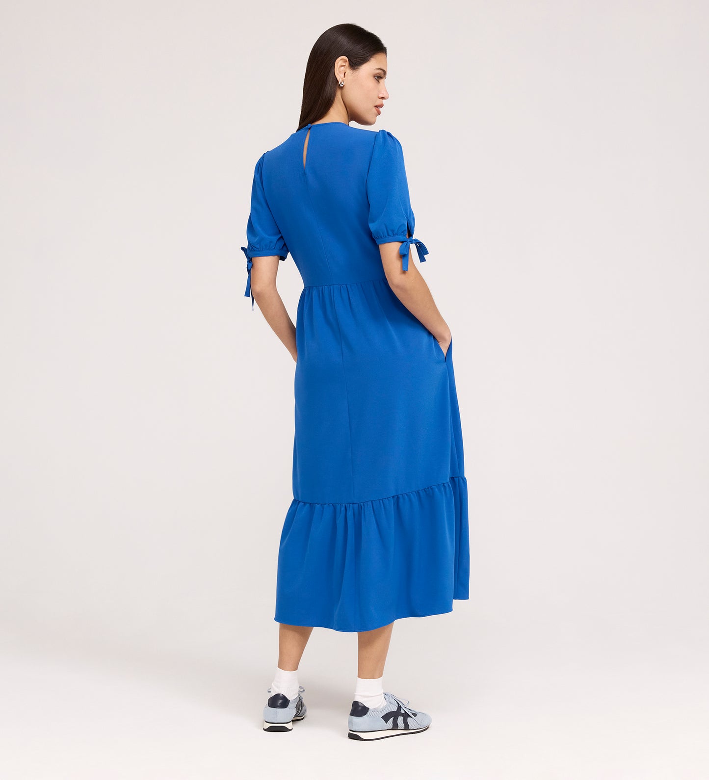 A woman with long brown hair faces away, wearing the Jessa Cobalt Crepe Midi Tiered Dress featuring short sleeves and tie details. She pairs it with white socks and sneakers against a plain, light background.