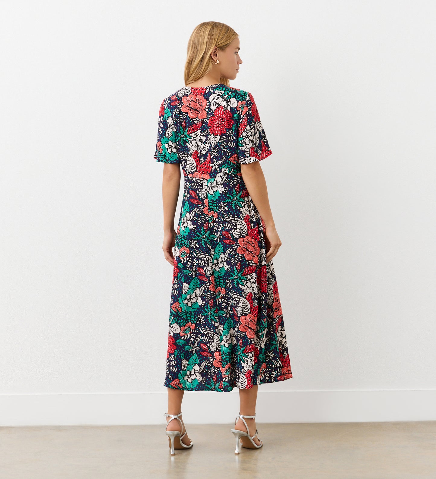 A woman with blonde hair wears the Josie Navy Floral Crepe Midi Dress, a calf-length piece featuring a bold red, white, and green floral print on dark crepe fabric, paired with light heeled sandals.