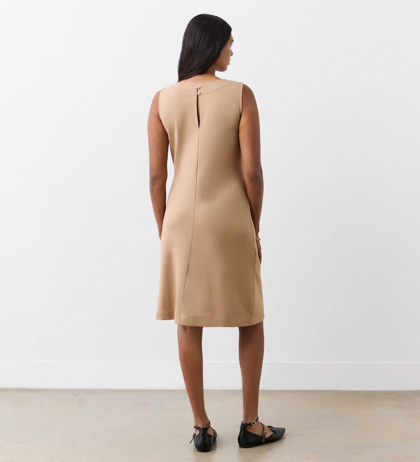 A woman with long dark hair faces away, wearing the Julianne Camel Ponte Jersey Midi Dress and black flat shoes, standing against a plain white wall and a light-colored floor.