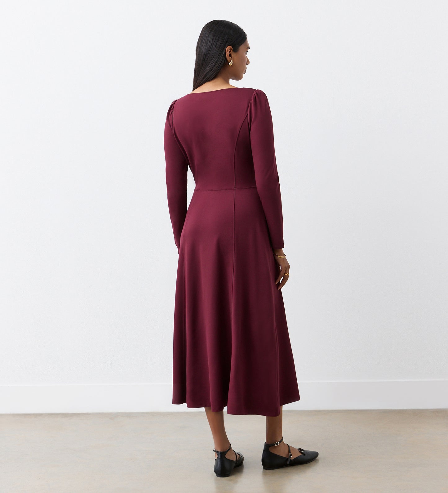 A woman stands indoors facing away, wearing the Kelly Burgundy Ponte Jersey Midi Dress—an essential with long sleeves, fitted waist, flared skirt, subtle seams—and pairs it with black flats against a plain white wall.