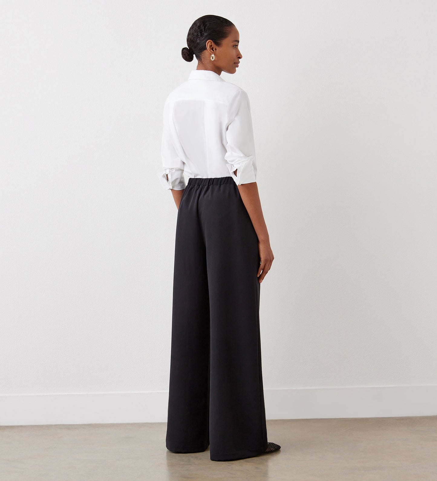 A woman stands facing away, wearing a white long-sleeve shirt with rolled-up sleeves and Nat Black Satin Back Crepe Wide Leg Trousers. Her hair is pulled back as she stands against a plain white wall and light floor.