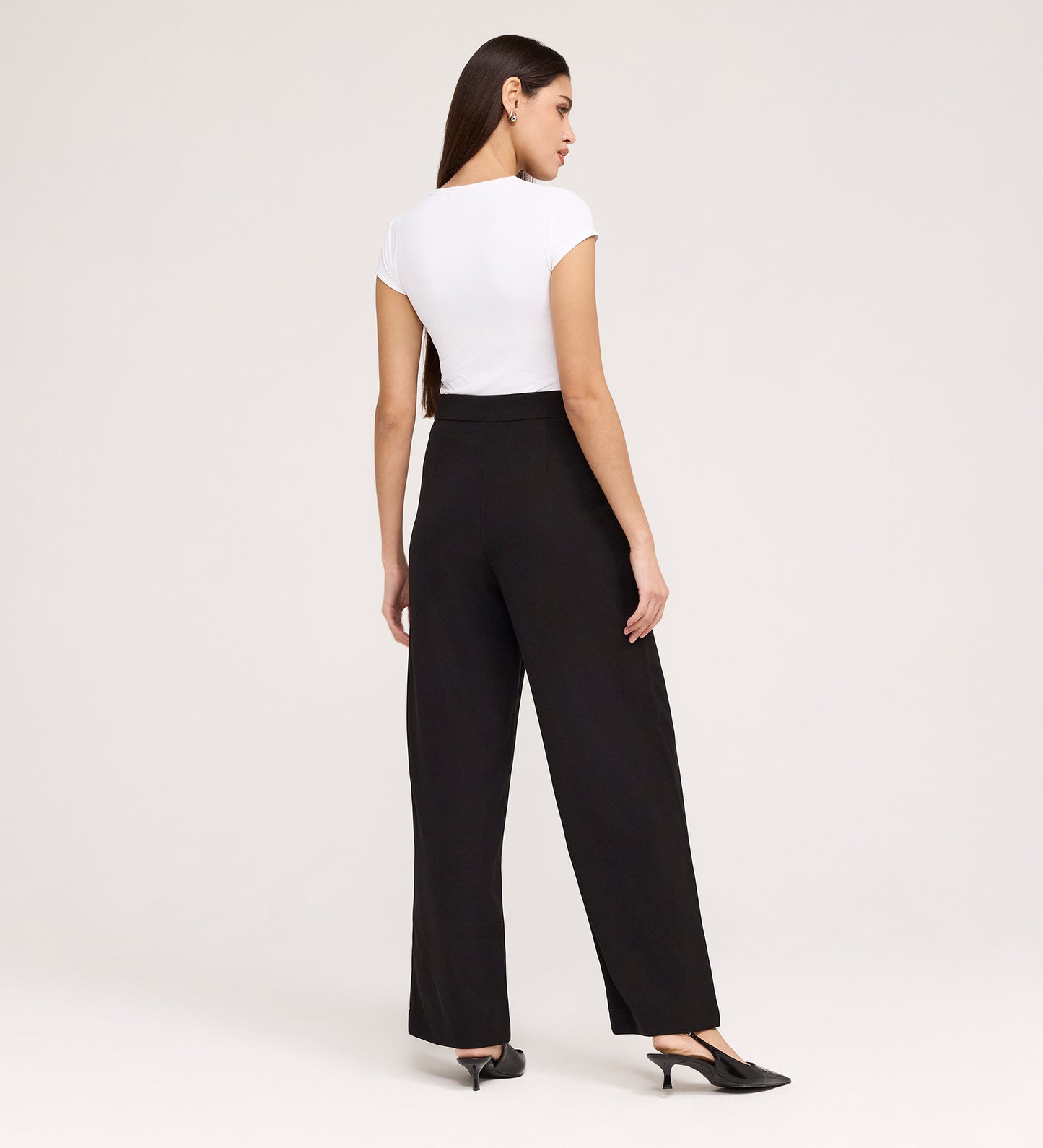 A woman, facing away, wears a fitted white short-sleeve top, black pointed heels, and Nellie Black Gaberdine Twill Wide Leg Trousers against a plain light background.