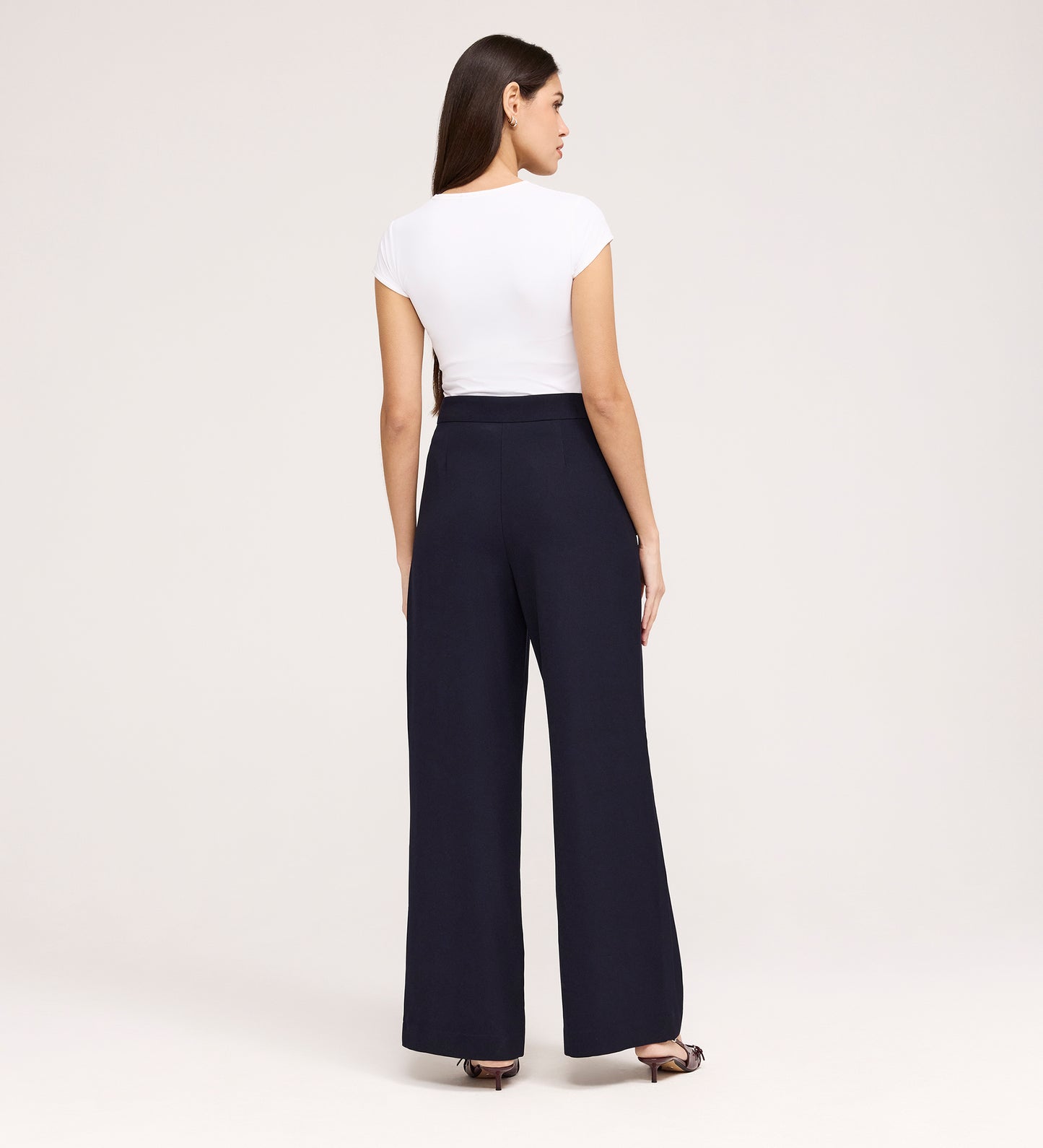 A woman faces away, wearing a fitted white top and Nellie Navy Gaberdine Twill Wide Leg Trousers with black open-toe heels against a plain light background—a true wardrobe staple.