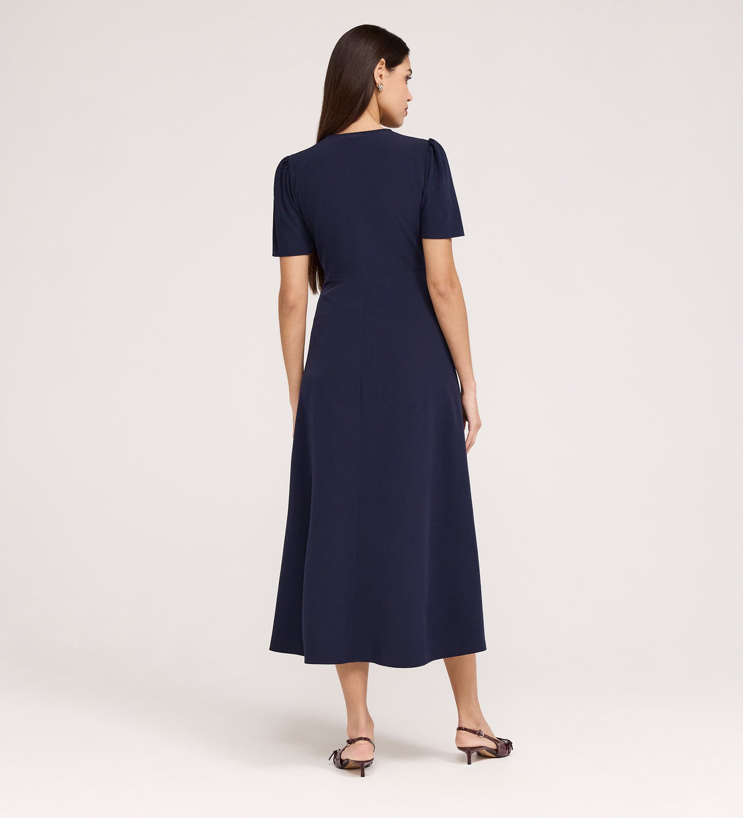 A woman with long brown hair stands facing away, wearing the Nieve Navy Crepe Midi Dress with a V-neckline and empire waist midi design, paired with open-toed heeled sandals against a plain light background.