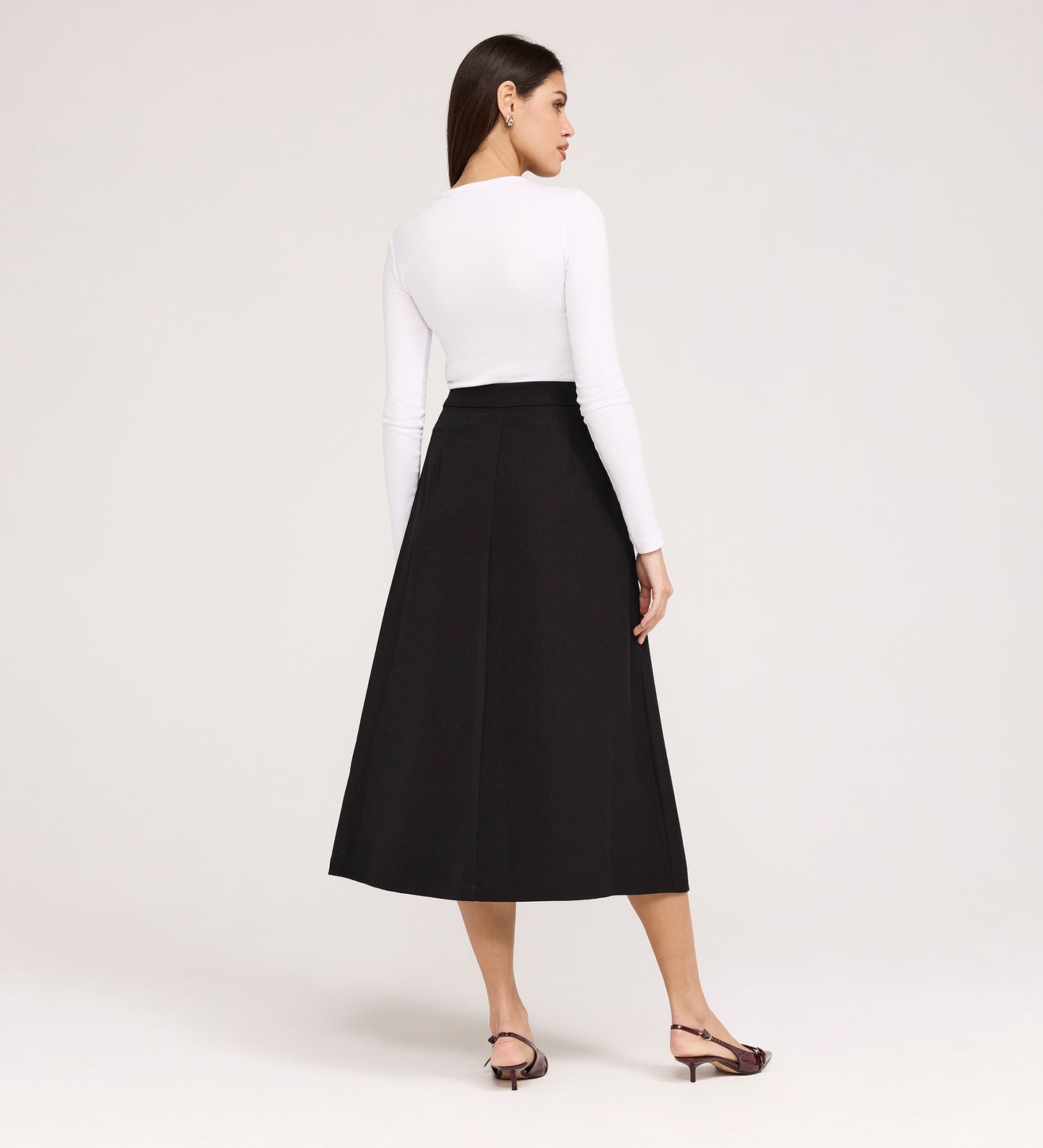 A woman faces away, wearing a long-sleeve white top paired with the Ramona Black Rich Crepe Midi Skirt and open-toed sandals. The background is plain and light-colored.