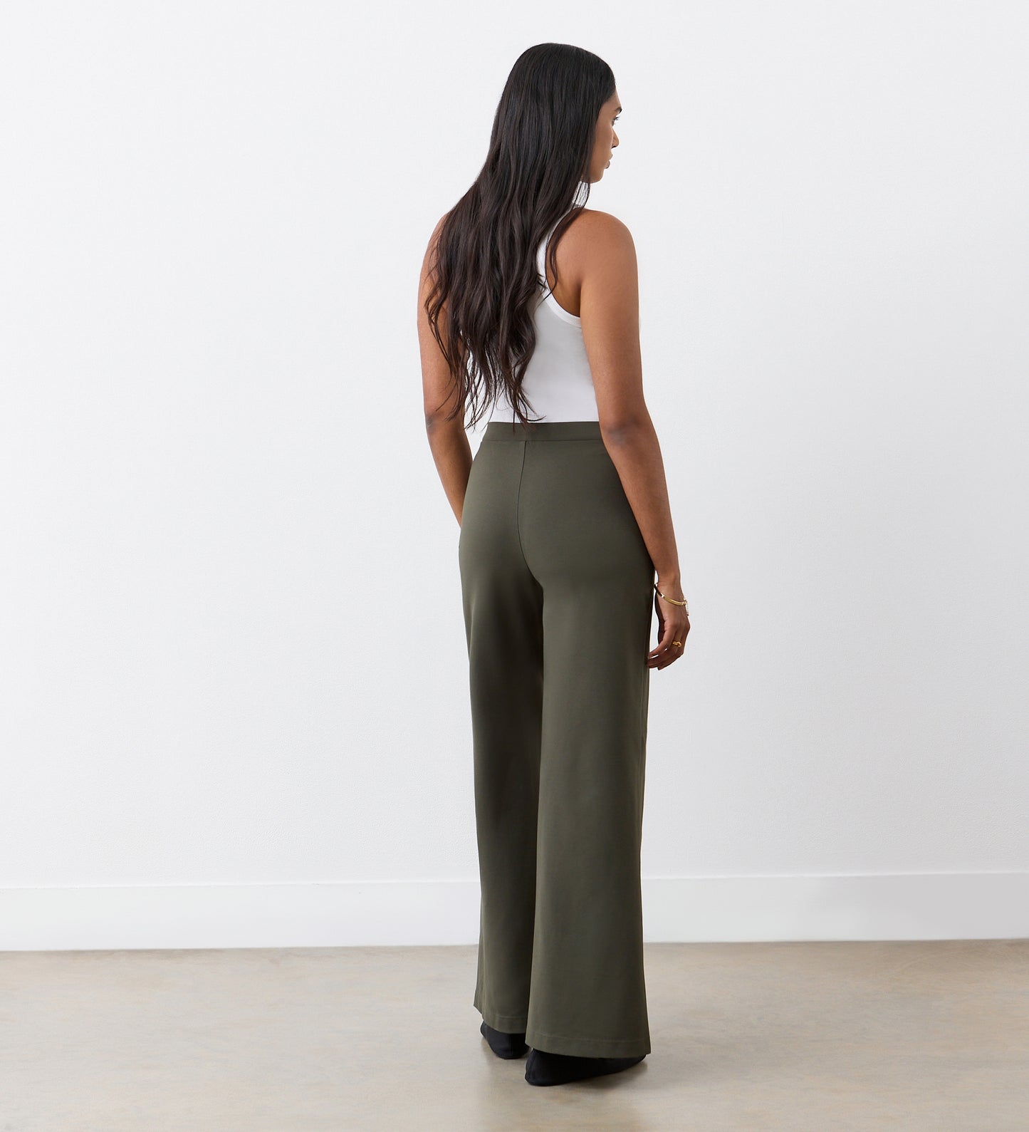 A woman with long dark hair, wearing a white sleeveless top and Reyna Khaki Ponte Jersey Wide Leg Trousers, stands facing away from the camera in a minimalist indoor setting.