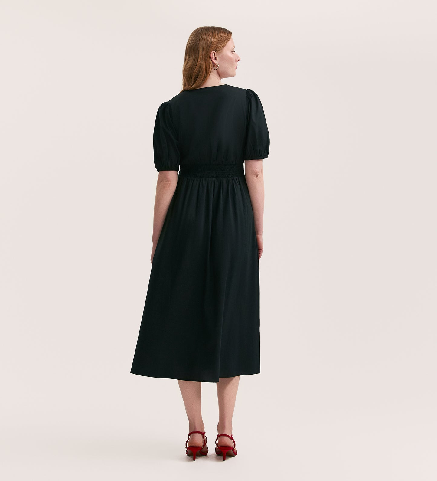 A woman with light brown hair faces away, wearing the Addison Black Cotton Midi Tea Dress featuring a v-neck, short puff sleeves, cinched waist, and seersucker fabric, styled with red heeled sandals against a plain light background.
