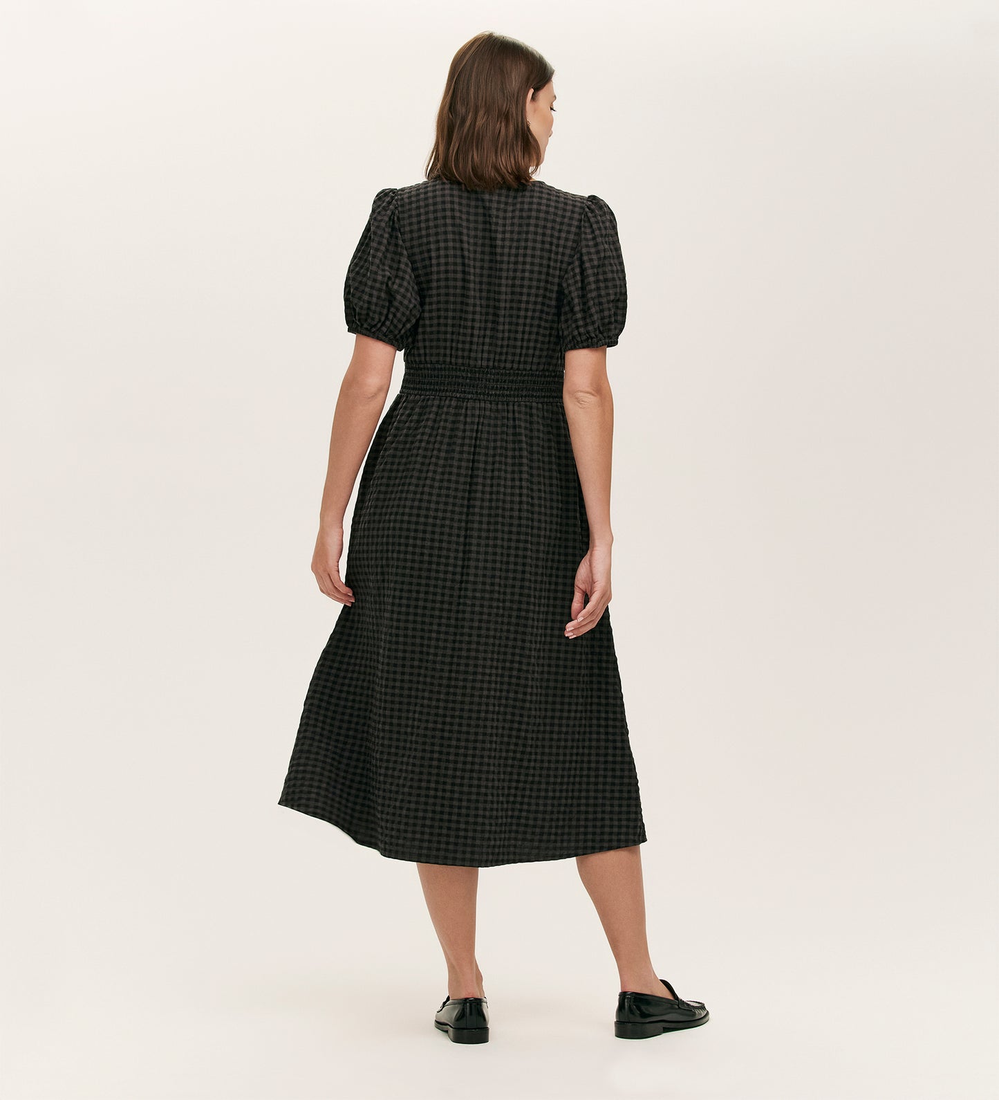 A woman, seen from behind, wears the Addison Black Seersucker Midi Tea Dress—featuring black and dark gray checks, cotton seersucker fabric, short puff sleeves—and black shoes against a plain light background.