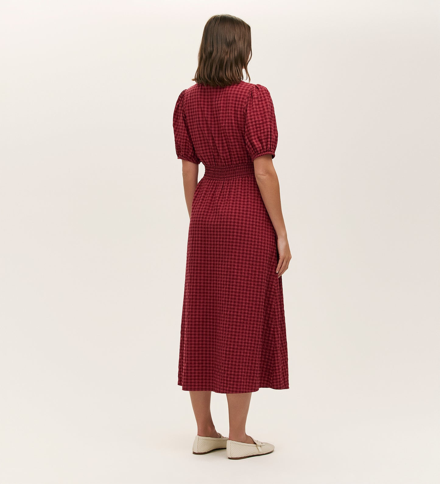 A woman with shoulder-length brown hair wears the Addison Burgundy Seersucker Midi Tea Dress, featuring a red and black checkered pattern and a fitted waist. She pairs it with white flats, standing against a plain light background.