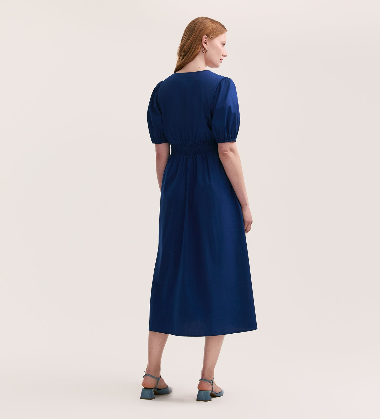 A woman with light brown hair faces away, wearing the Addison Indigo Cotton Midi Tea Dress with short puffed sleeves and blue heeled sandals, set against a plain light background.
