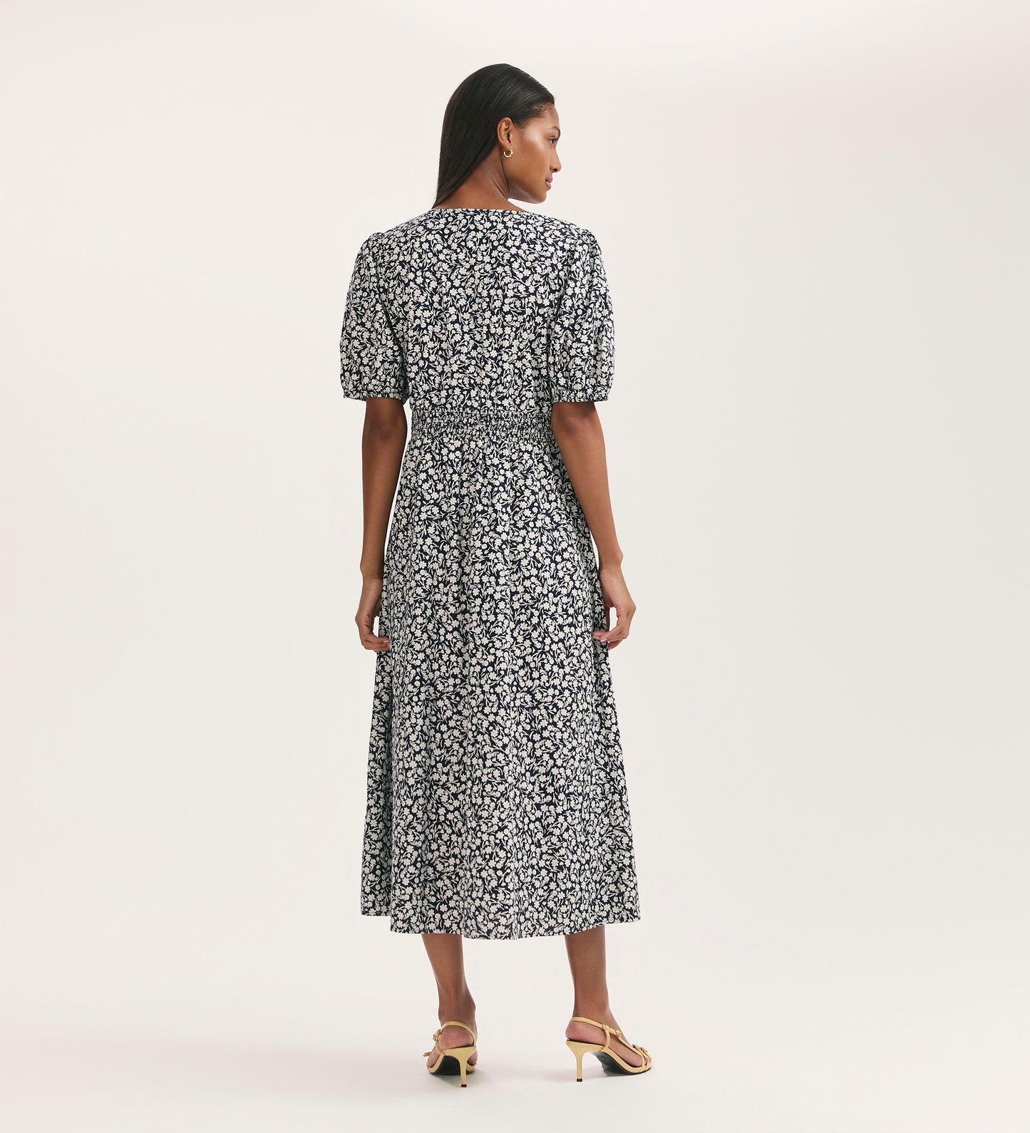 A woman faces away, wearing the Addison Navy Flower Cotton Button Through Midi Dress with white floral patterns, short puff sleeves, and beige heeled sandals against a plain light background.
