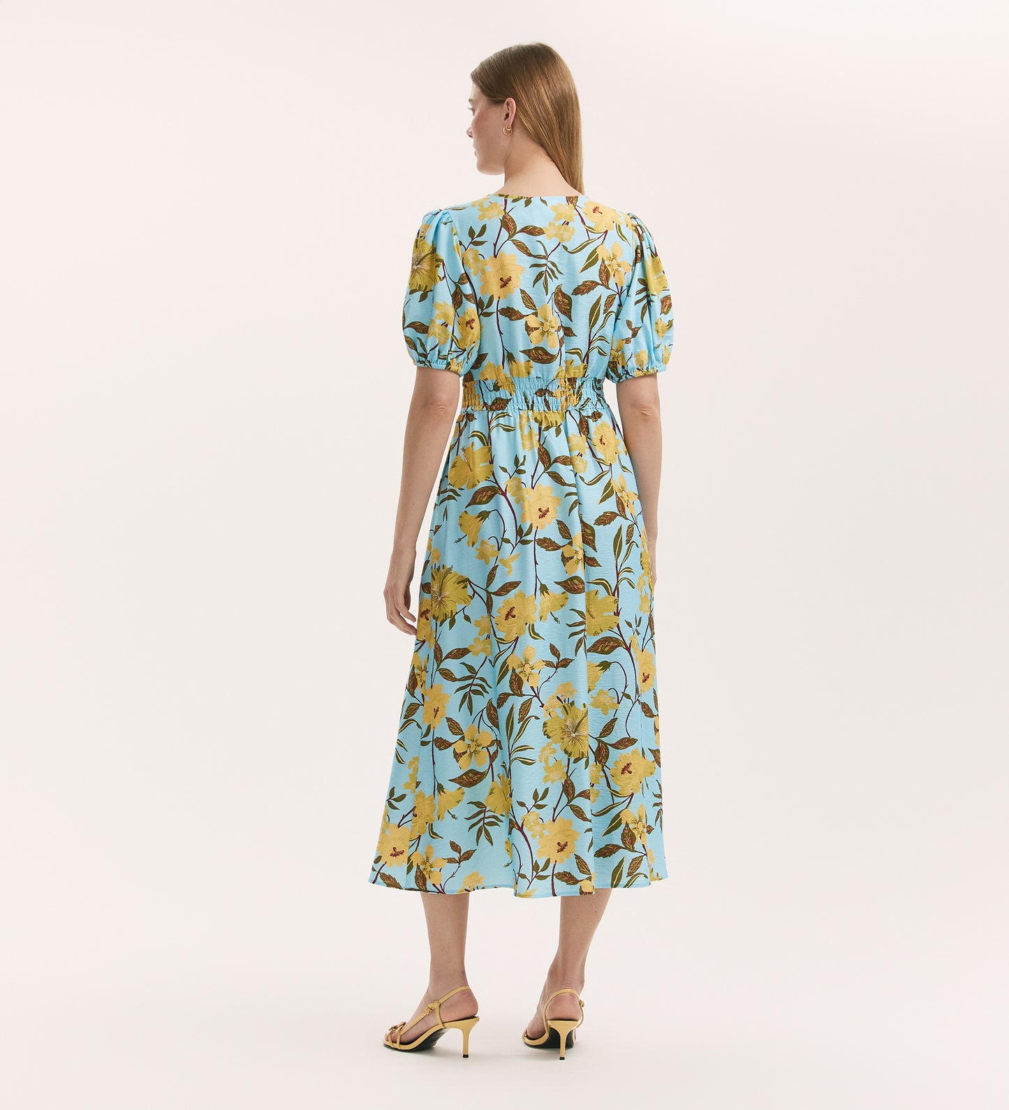 A woman with straight light brown hair faces away, wearing the Addison Blue Floral Crepe Button Through Midi Dress featuring yellow floral patterns and short puff sleeves. She pairs it with yellow heeled sandals against a plain white background.