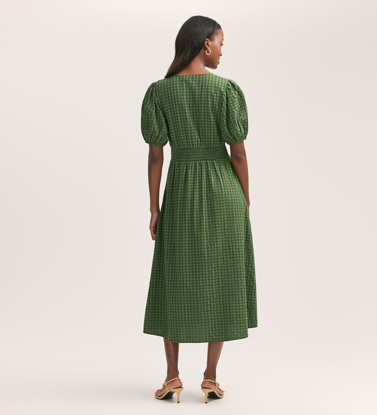 A woman stands facing away, wearing the Addison Khaki Gingham Seersucker Midi Dress with short puff sleeves and a fitted waist. Her long dark hair cascades over the textured fabric as she styles it with heeled sandals against a light background.