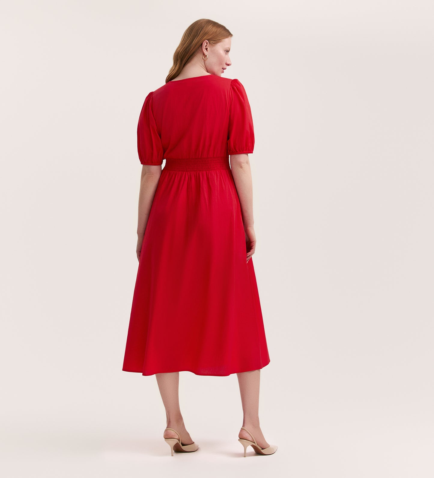 A woman with light skin and long, straight blonde hair wears the Addison Red Cotton Midi Tea Dress with short puff sleeves and beige heels, standing against a plain light background, facing away from the camera.