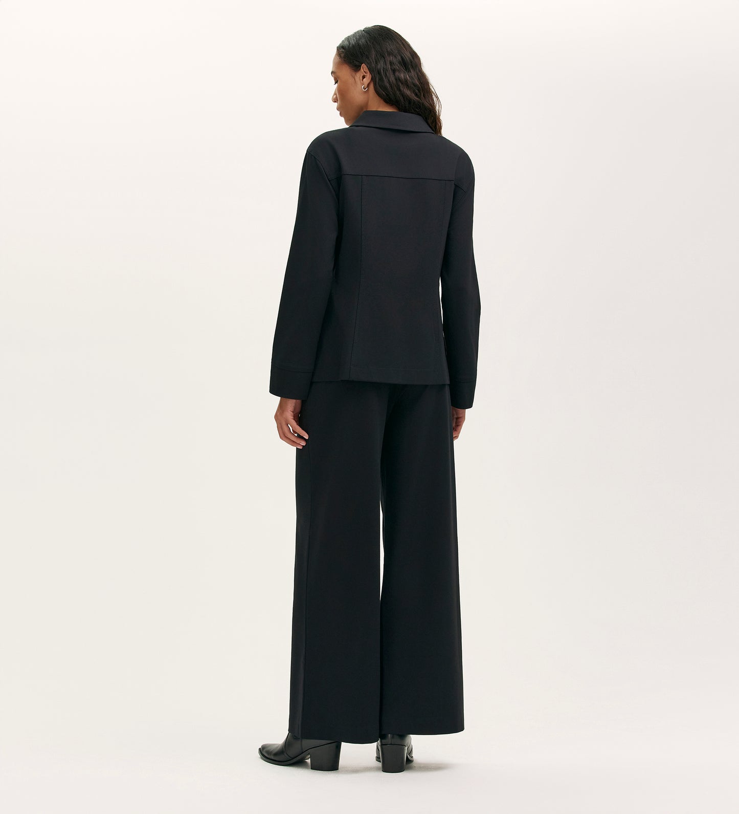 A woman faces away, wearing the Agness Black Ponte Jersey Button Down Jacket with matching wide-leg pants and black shoes, elegantly styled against a plain light background.