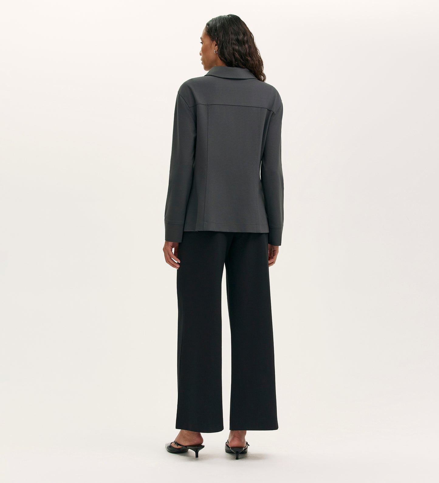 A woman stands facing away from the camera, wearing the Agness Charcoal Ponte Jersey Button Down Jacket over a long-sleeve dark gray top, wide-leg black pants, and black heeled sandals against a plain light background.