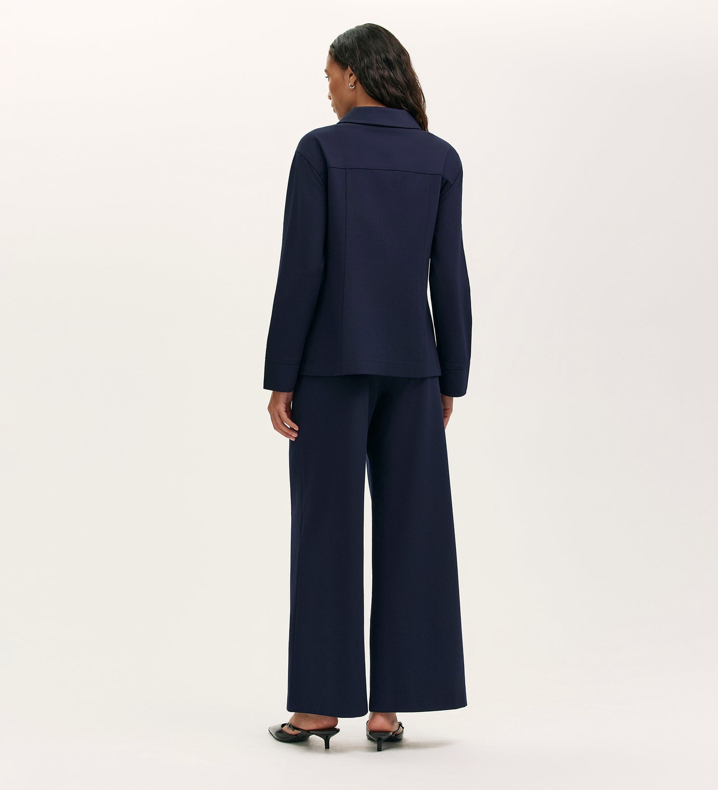A woman faces away from the camera wearing the Agness Navy Ponte Jersey Button Down Jacket with wide-leg pants and heeled shoes, set against a plain, light background.