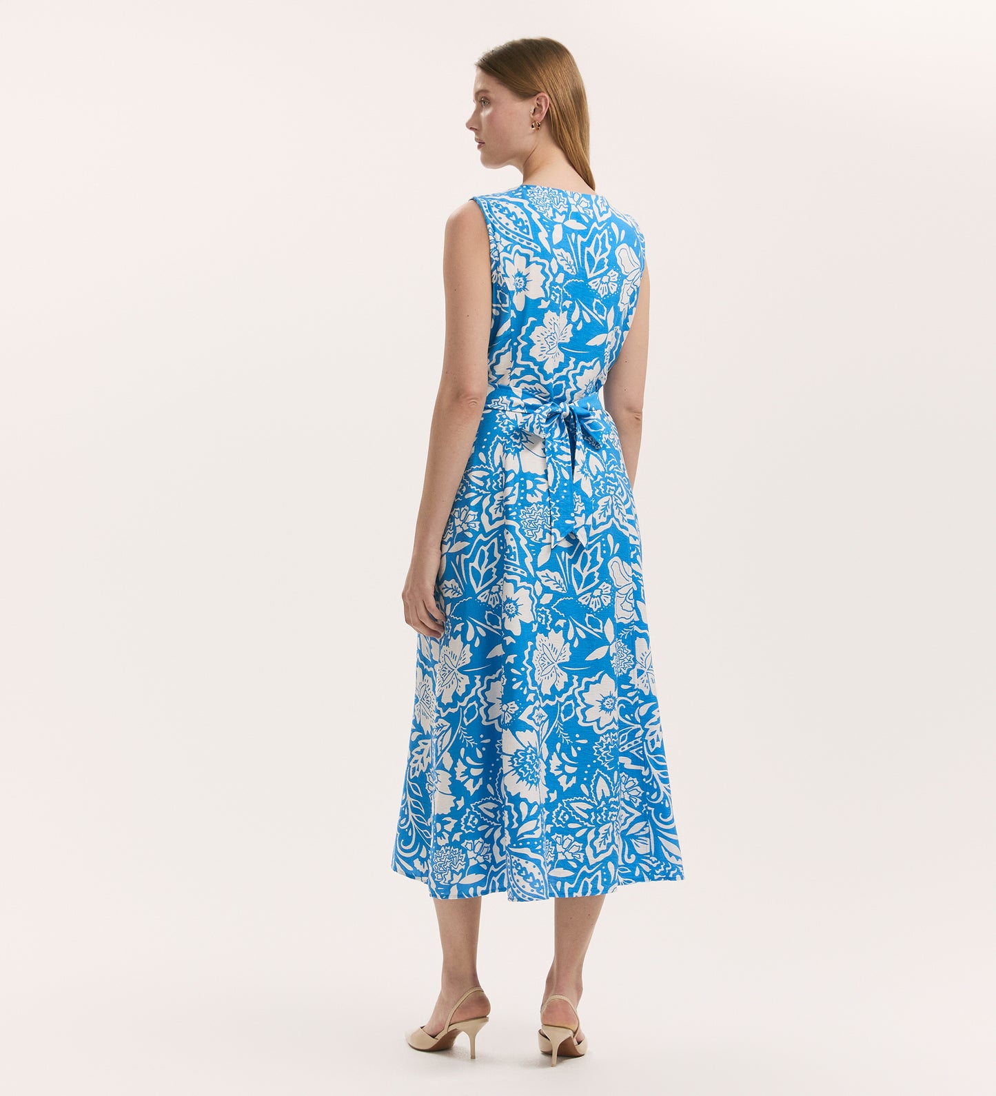 A woman with light skin and straight hair faces away, wearing the Aimee Blue Flower Button Through Sleeveless Midi Dress and beige heeled shoes, set against a plain light background.