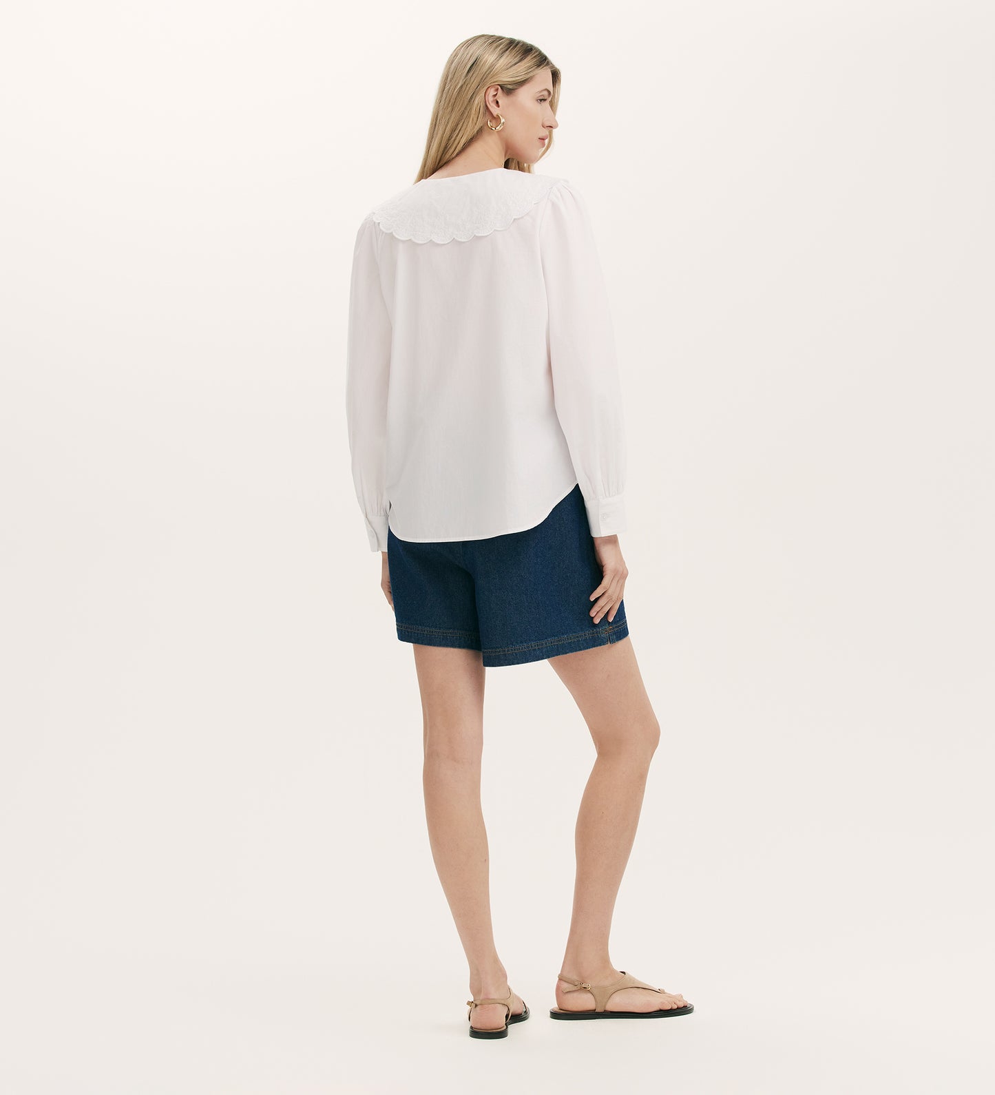 A woman with straight blonde hair stands against a plain light background, wearing the Ansley White Cotton Shirt—a relaxed fit long-sleeve blouse with an embroidered Peter Pan collar—paired with dark denim shorts and beige sandals.