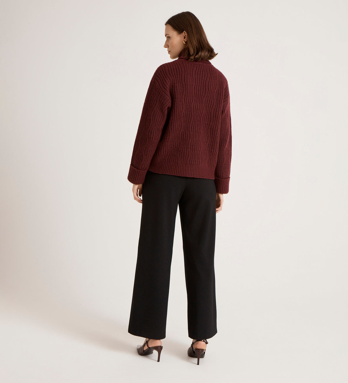 A woman stands facing away, wearing the Albie Burgundy Roll Neck Jumper, paired with black wide-leg pants and black heeled sandals against a plain light background.