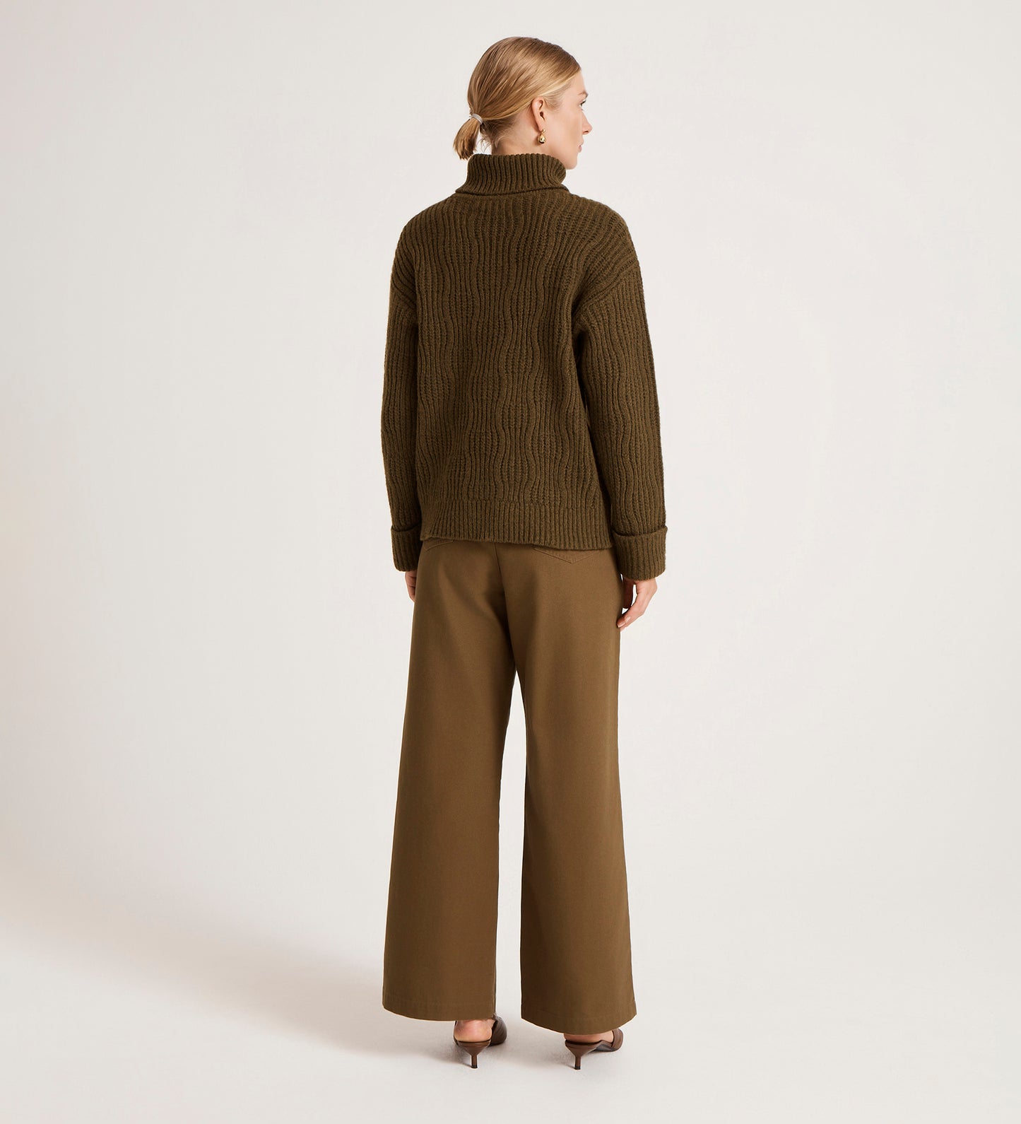 A woman faces away against a plain light background, wearing the Albie Khaki Roll Neck Jumper and wide-leg brown trousers paired with heeled shoes.