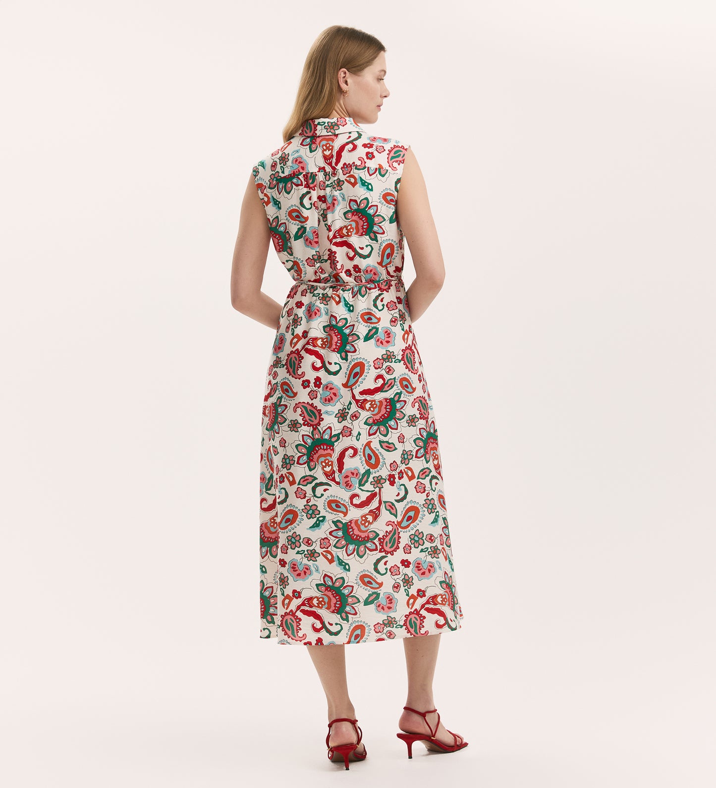 A woman with light hair, seen from behind, models the Alida Ivory Paisley Crepe Sleeveless Midi Dress featuring an elastic waist and red-green paisley floral print. She pairs it with red heeled sandals against a plain, light background.