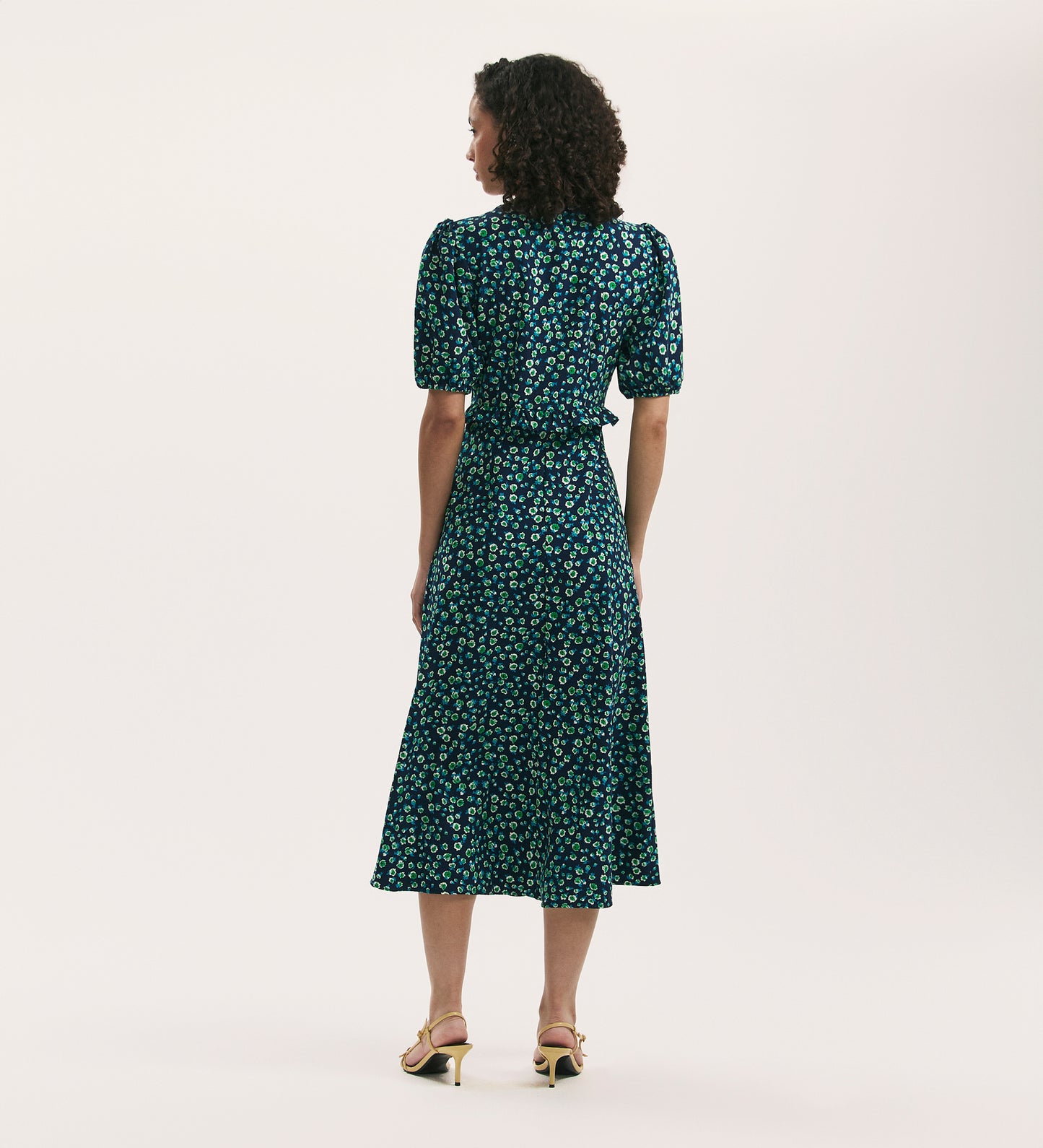A woman, seen from behind, wears the Anika Navy Ditsy Crepe Midi Tea Dress with puffed short sleeves in crease-free fabric, paired with yellow heeled sandals, standing against a light background.