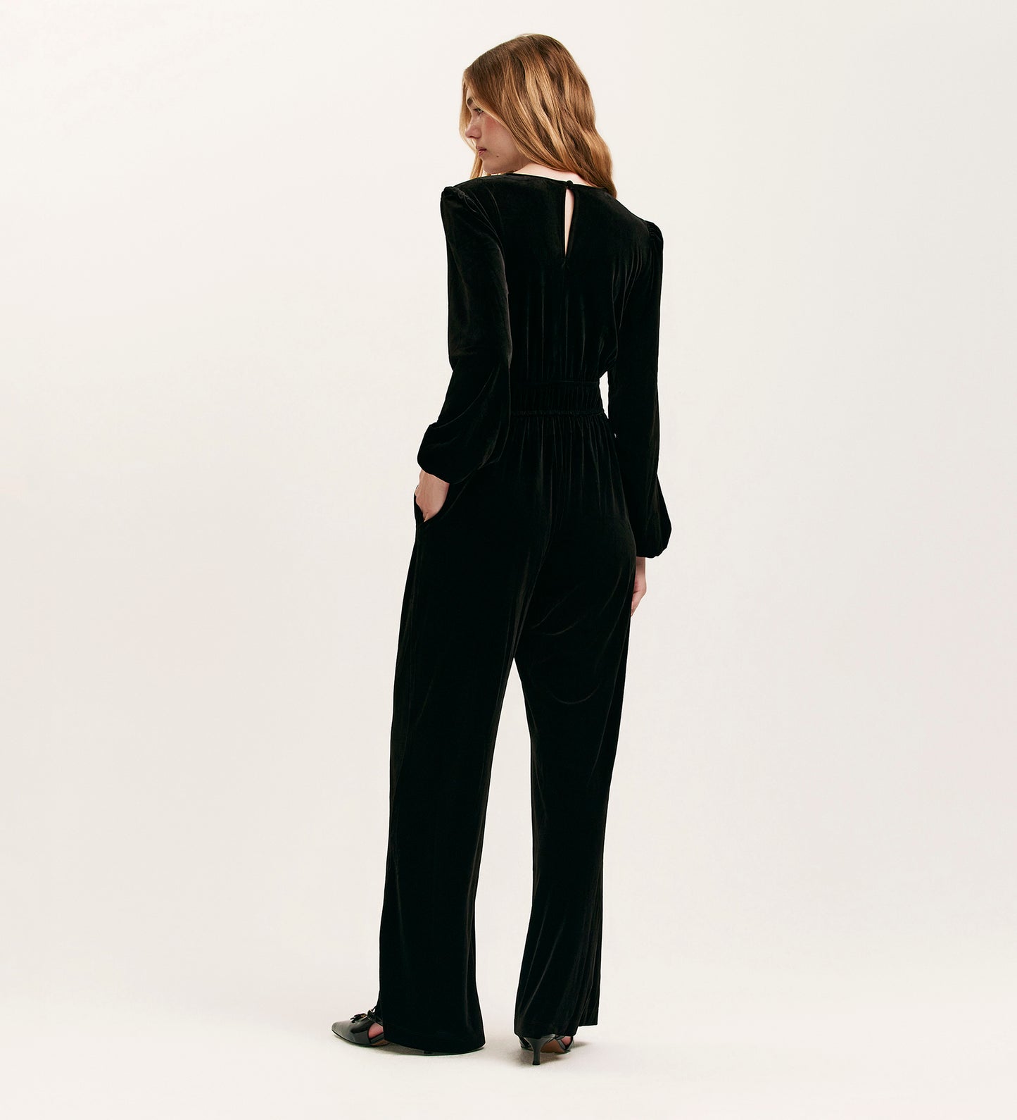 A woman stands facing away from the camera in the Anna Black Velvet Jumpsuit, featuring long sleeves and wide legs, paired with black open-toed heels for a chic, relaxed-fit look against a plain white background.