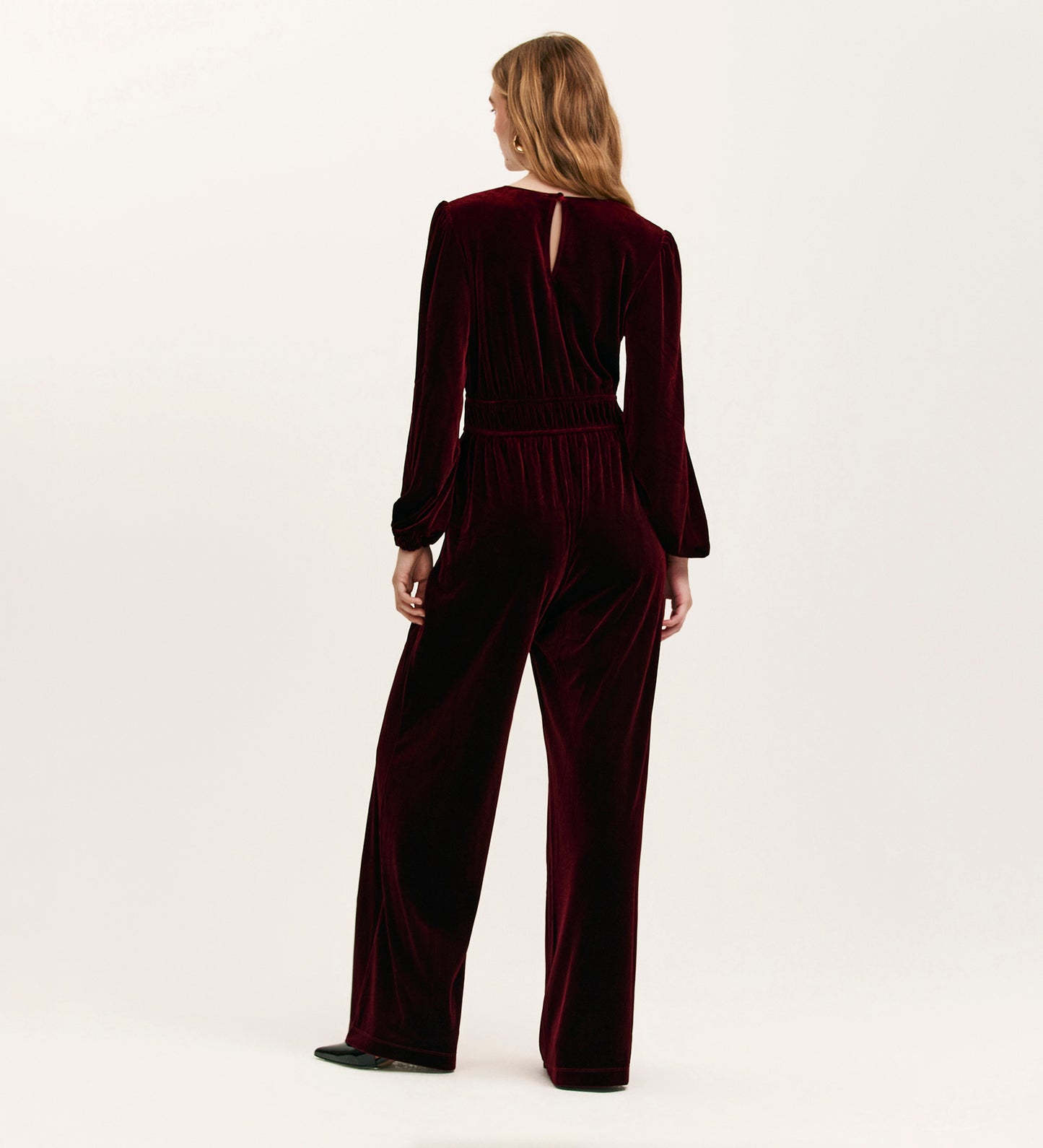 A woman with light brown wavy hair stands against a white background, wearing the Anna Burgundy Velvet Jumpsuit— a relaxed-fit, wide-leg design in dark burgundy jersey velvet with an upper-back keyhole opening.